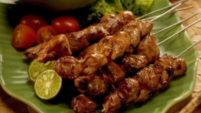 Sepiring sate kambing dengan tusuk sate disusun rapi di atas piring hijau, dilengkapi potongan jeruk nipis, tomat ceri, daun selada, dan semangkuk kuah gulai berwarna kuning di latar belakang.