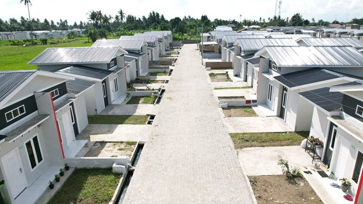 Kompleks perumahan dengan deretan rumah tipe kecil beratap seng berjajar rapi di kedua sisi jalan paving, dikelilingi area hijau dan pohon kelapa di kejauhan.