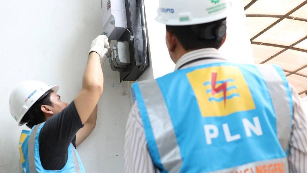 Dua petugas PLN mengenakan helm dan rompi keselamatan sedang memeriksa dan memasang meteran listrik di dinding bangunan.