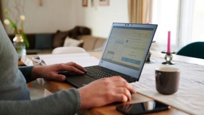 Seorang pria sedang bekerja menggunakan laptop di atas meja kayu dengan latar ruang tamu, terdapat secangkir kopi, smartphone, dan beberapa peralatan meja lainnya.