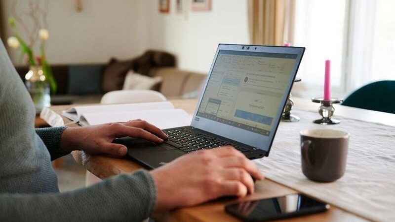 Seorang pria sedang bekerja menggunakan laptop di atas meja kayu dengan latar ruang tamu, terdapat secangkir kopi, smartphone, dan beberapa peralatan meja lainnya.