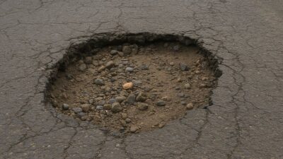Lubang besar di permukaan jalan aspal dengan batu dan tanah terlihat di dalamnya, berada di tengah jalur lalu lintas jalan raya yang sepi dengan pepohonan di sisi kanan dan kiri.