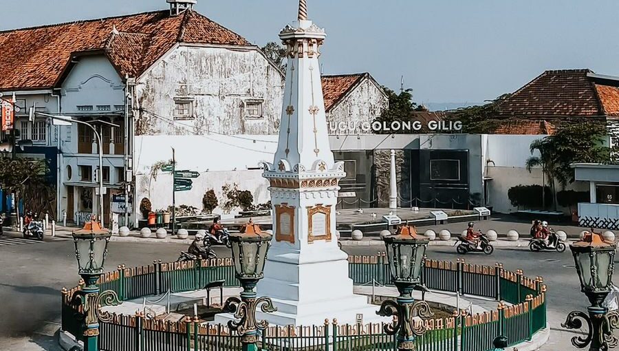 Tugu Jogja yang berwarna putih berdiri di tengah perempatan jalan dengan lalu lintas sepeda motor yang melintas, berlatar belakang langit biru cerah dan bangunan bergaya kolonial di Yogyakarta.